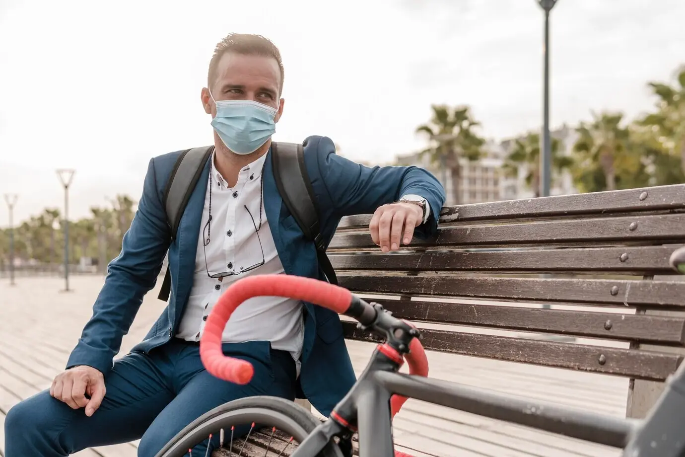 A man wearing a face mask sits on a bench beside his bicycle outdoors.